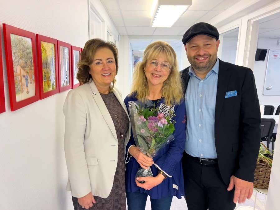 Tre glada personer i ett ljust kontor. Pristagaren står i mitten med blommor i handen.