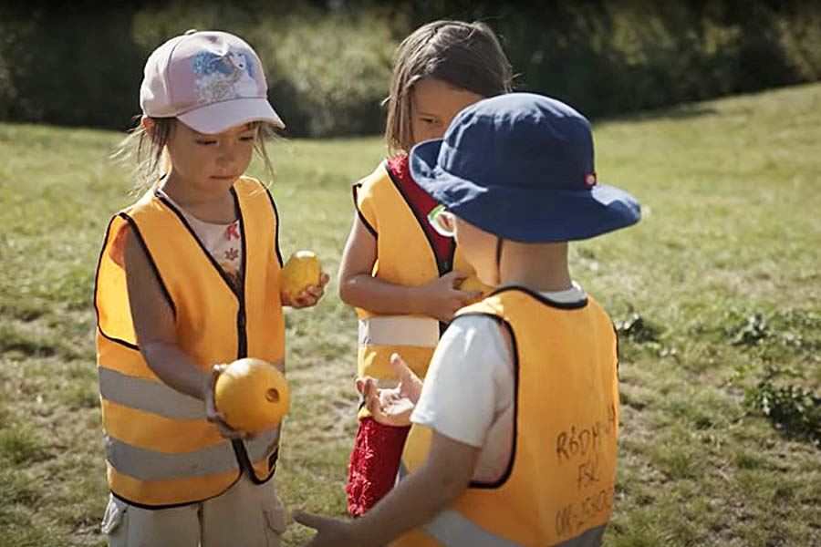 Tre barn på förskola i gula västar leker i naturen. 