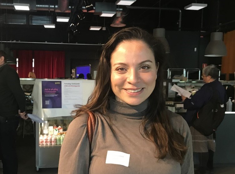 Kvinna i brun tröja står i en inomhuslokal och tittar in i kameran
