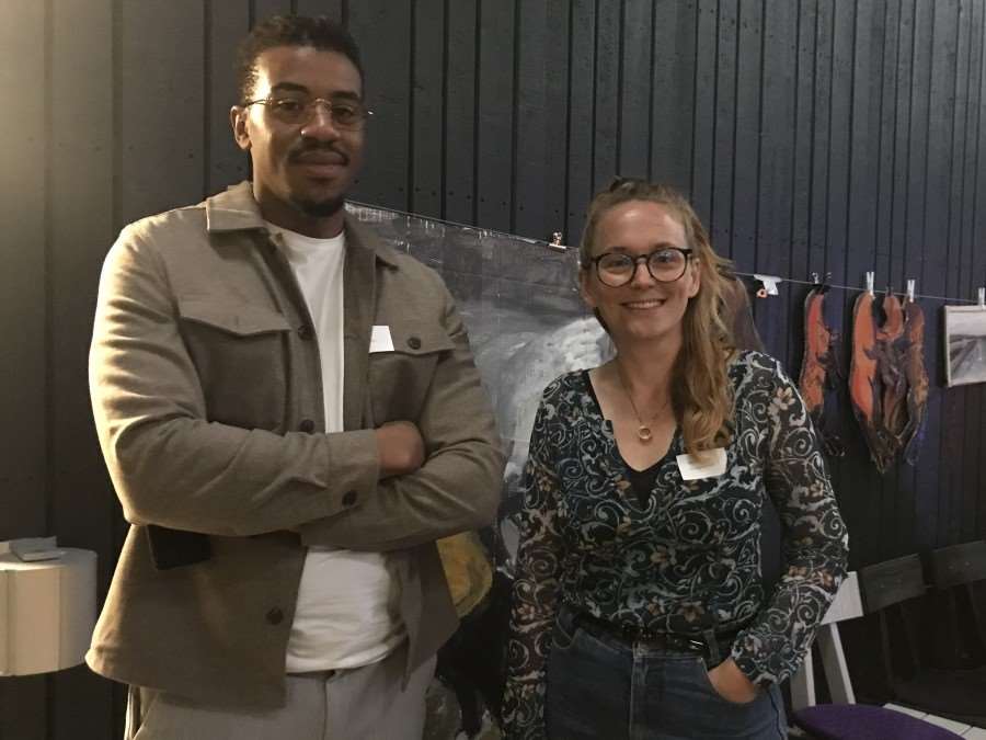 En man och en kvinna står mot en svart vägg med tavlor och tittar in i kameran