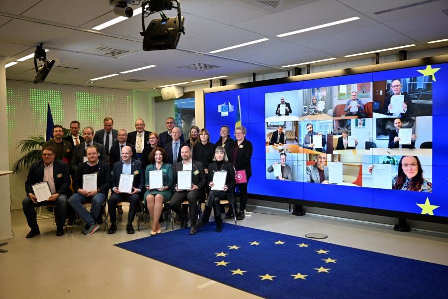 Gruppbild på personer som både sitter på stolar och personer som är med på skärm på väggen, alla håller i ett påskrivet kontrakt