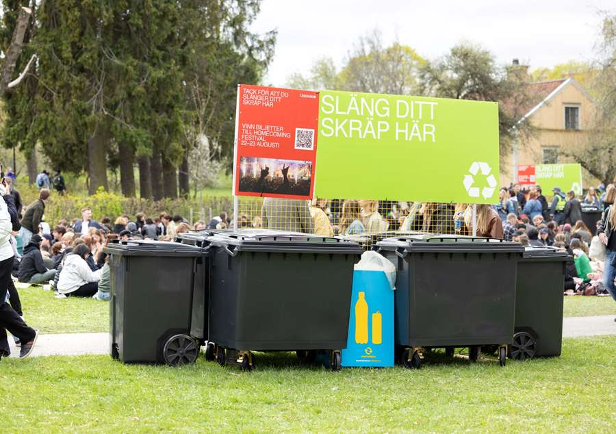 Sopcontainers vid Ekonomikumparken i Uppsala. Folk firar valborg i bakgrunden.