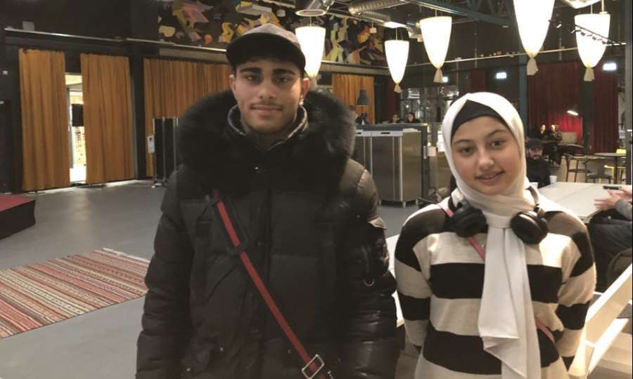 En tonårspojke och en tonårsflicka står i ett kafé och tittar in i kameran