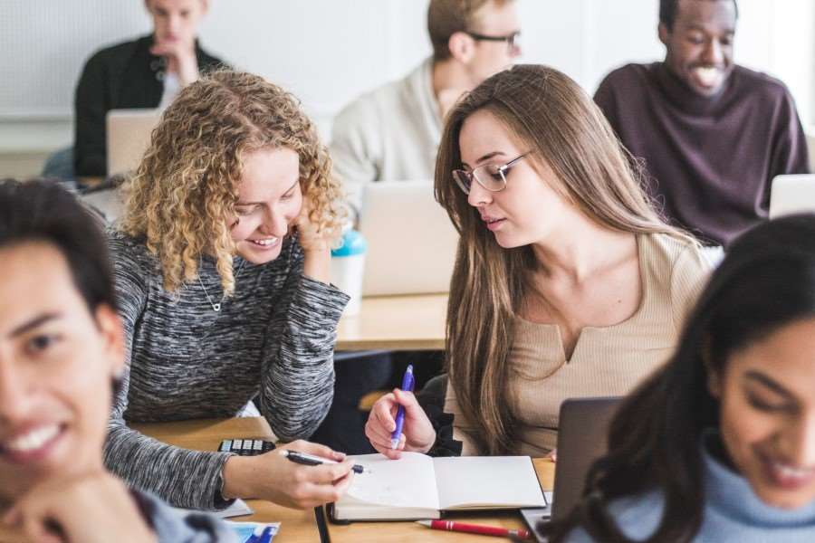 Två kvinnor sitter i ett klassrum bland studenter och tittar i en bok