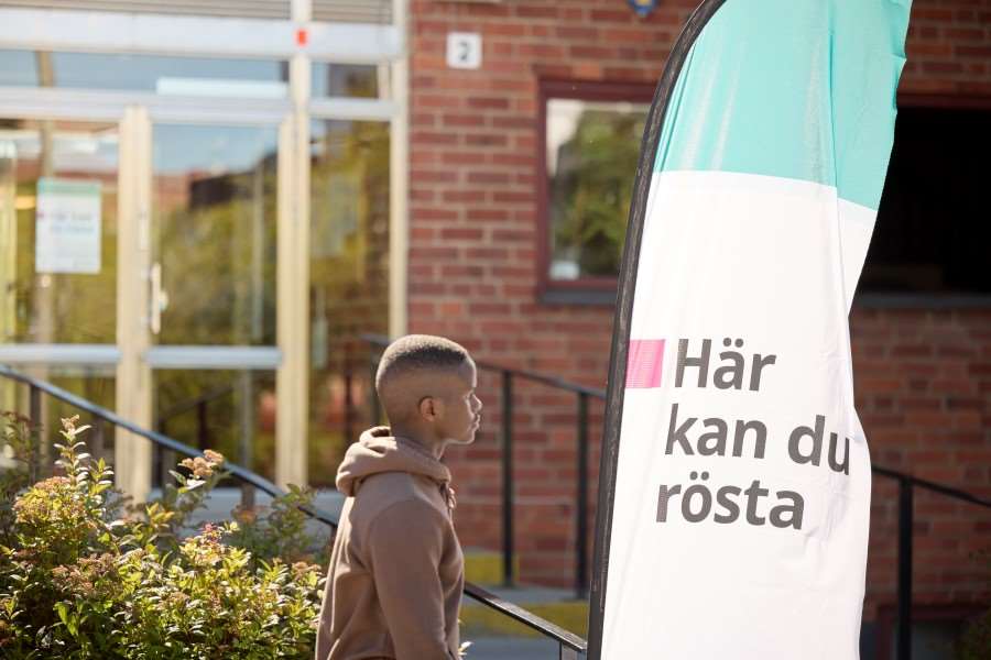 En person går framför en byggnad och en flagga som det står "Rösta här" på.