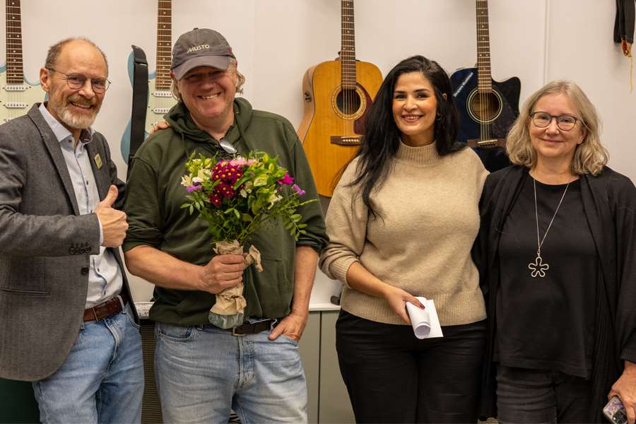 Fyra personer ler mot kameran, en håller en bukett i handen, gitarrer hänger på väggen bakom