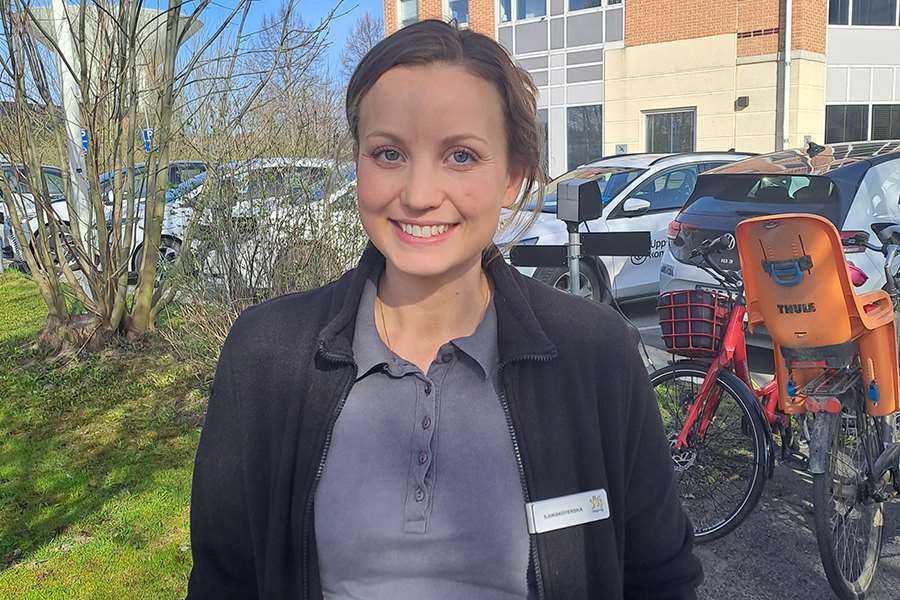 Kvinna framför en tegelbyggnad och några cyklar. Hon har på sig en grå kofta och en pikétröja