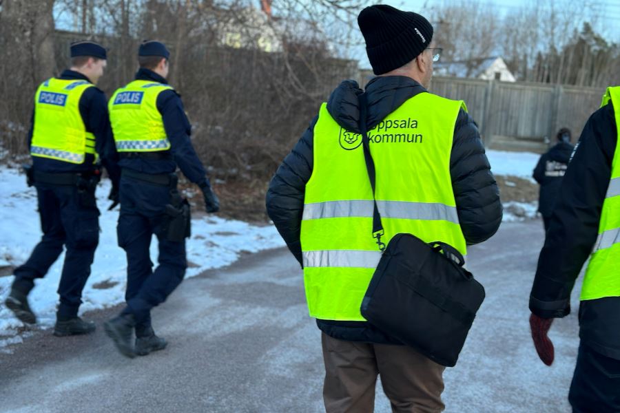 Tv&aring; poliser och en man med gul v&auml;st med Uppsala kommuns logotyp promenerar, de &auml;r fotograferade bakifr&aring;n.