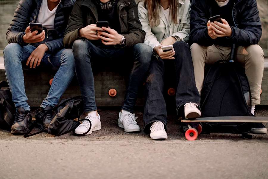 Fyra ungdomar sitter på en bänk och tittar i sina mobiler knän och händer syns