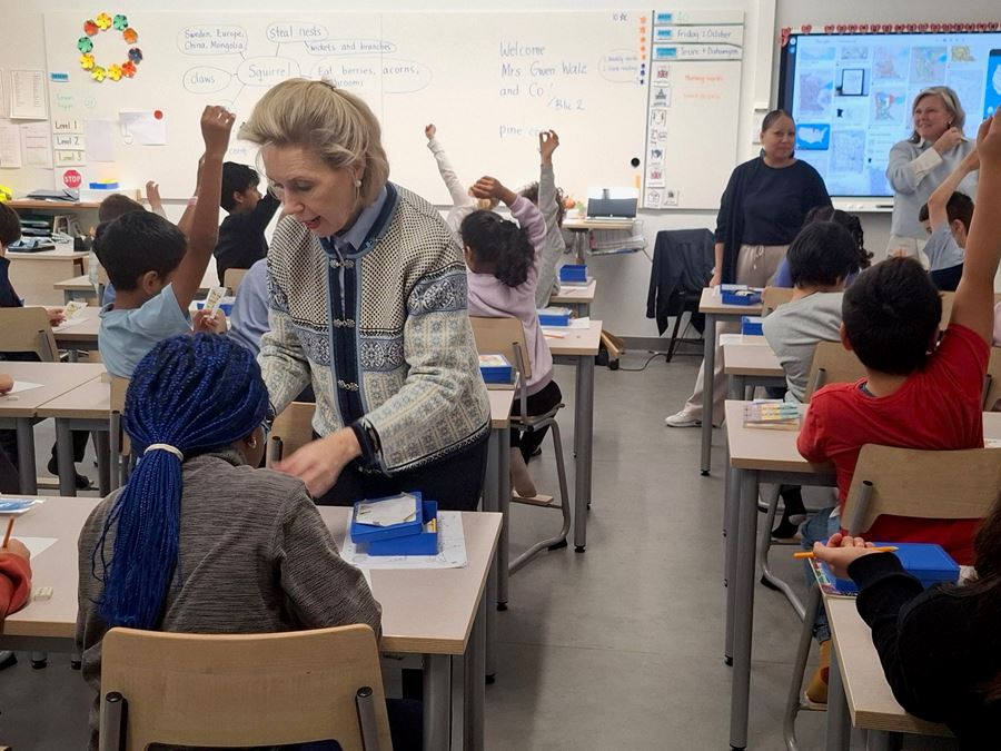 En äldre kvinna står mitt i ett klassrum där elever sitter vid bänkarna.