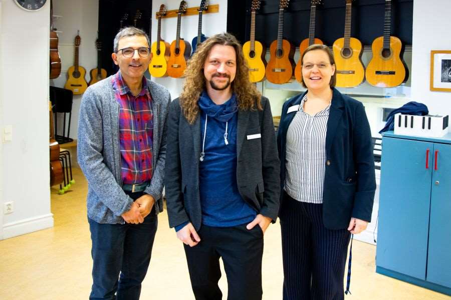 Tre personer ler mot fotografen, gitarrer hänger på väggen i bakgrunden