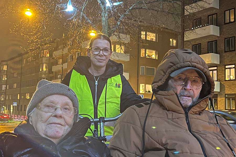 En äldre dam och en äldre man sitter i en lådcykel. Bakom dem sitter en kvinna i reflexväst och bakom henne syns en ljusinstallation