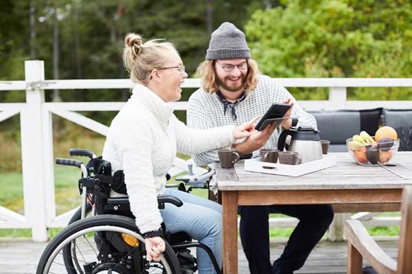 Manlig vårdgivare och funktionshindrad kvinna sitter vid bord på altan och använder surfplatta tillsammans.