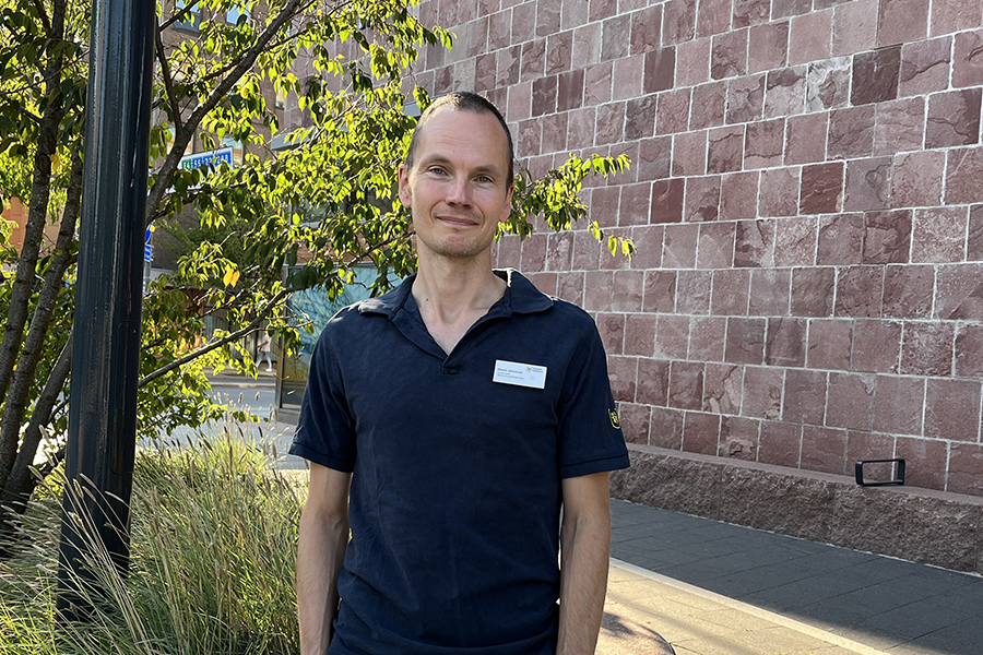 En man vid en tegelvägg och gröna träd. Han bär kommunens kläder.