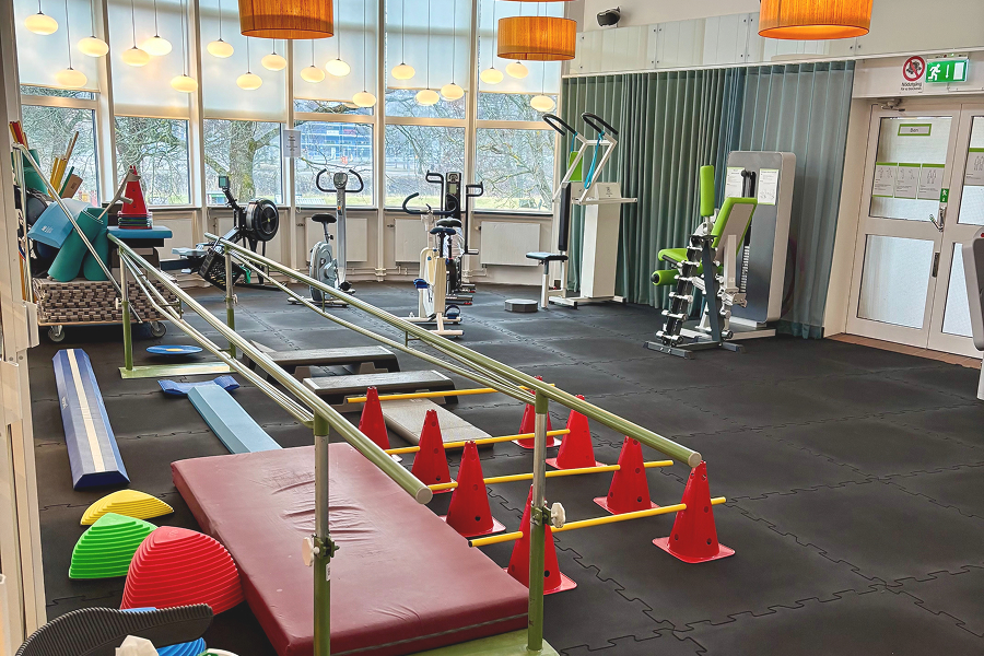 Ett ljust träningsrum med olika gymredskap, balansutrustning och en gångbana med räcken och koner på ett mjukt golv.