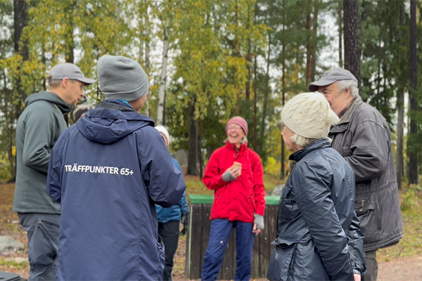 Fem personer står och pratar i en skog. En person har en jacka där det står Träffpunkt 65+