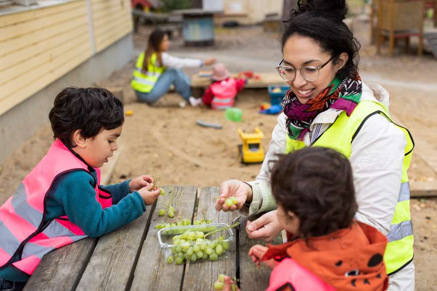 En förskolelärare sitter vid ett bord med två barn  och äter vindruvor.