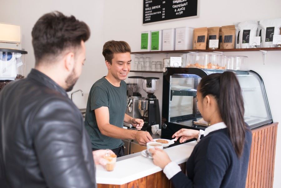 En ung person serverar kaffe till en kund efter en caf&eacute;disk.