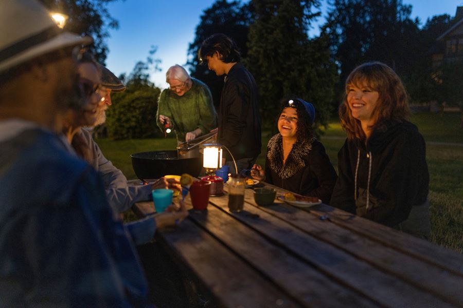 Personer sitter utomhus vid ett bord i skymningen. De har en batteridriven lykta