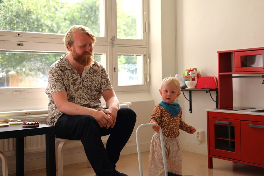 En pappa sitter i en soffa och ett barn står bredvid med en liten vagn.