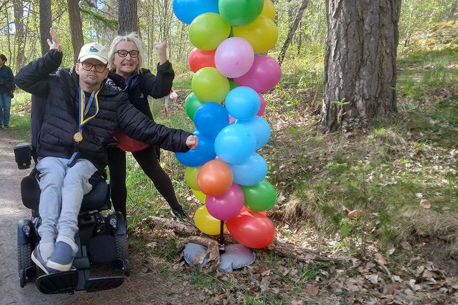 En man i rullstol bredvi en kvinna i en skog. Ballonger syns också.