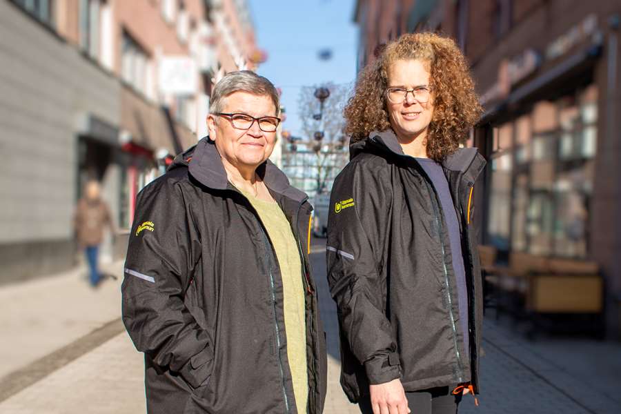 Två leende kvinnor, klädda i jackor med Uppsala kommuns logotyp, står i solen .