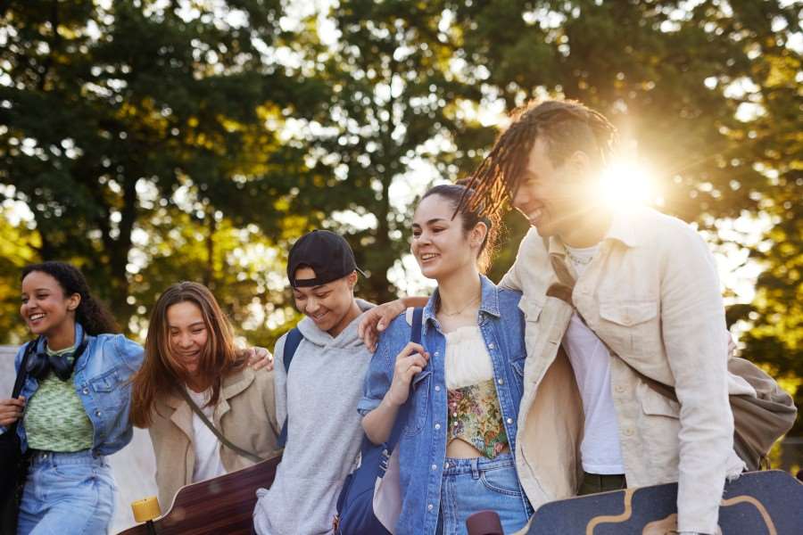 En grupp på fem ungdomar går tillsammans utomhus i en park med grönskande träd i bakgrunden. Solen skiner lågt bakom dem och skapar motljus. Personerna har armarna om varandras axlar och några bär skateboards. De är klädda i vardagliga kläder som jeans, hoodie och jackor, och rör sig tillsammans längs en gång.