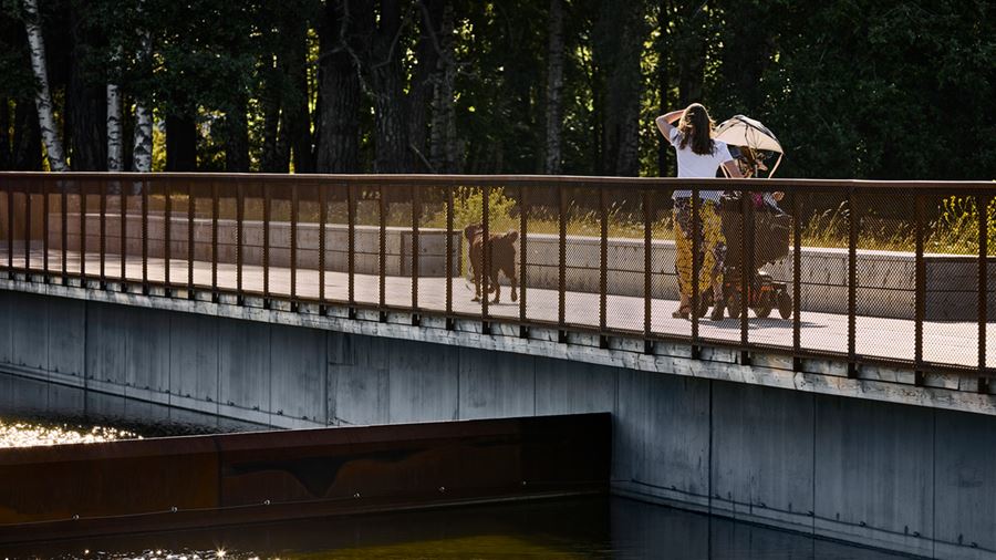Kvinna med barnvagn och en hund promenerar på bryggan runt dagvattendammen.