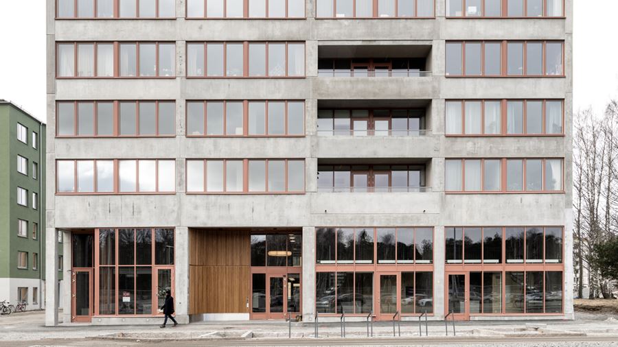 Raw, exteriörbild på flerbostadshus med ljusgrå betongfasad och brunröda fönsterkarmar.
