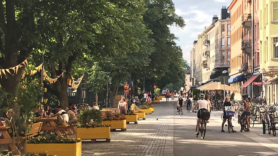 Sommargatan p&#229; &#214;stra &#197;gatan mellan S:t Olofsgatan och Bang&#229;rdsgatan.  Mellan tr&#228;den h&#228;nger vimplar i gult och orange. L&#228;ngs gatan finns bloml&#229;dor och b&#228;nkar i gult. D&#228;r sitter m&#228;nniskor och umg&#229;s. P&#229; gatan bredvid cyklar och g&#229;r m&#228;nniskor.