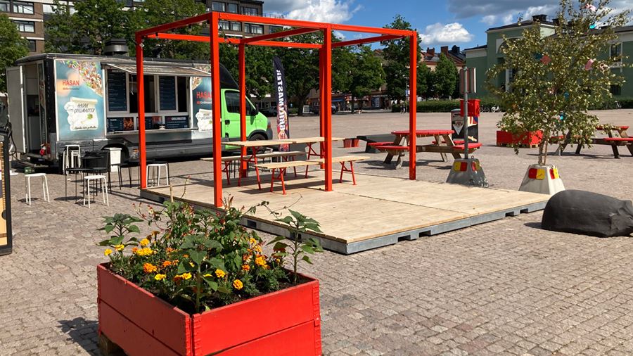 Sommartorg p&#229; Vaksala torg. Blomsterplanteringar i r&#246;da l&#229;dor och r&#246;dm&#229;lade sittplatser. Intill sommartorget finns en  matvagn d&#228;r det g&#229;r att k&#246;pa mat.