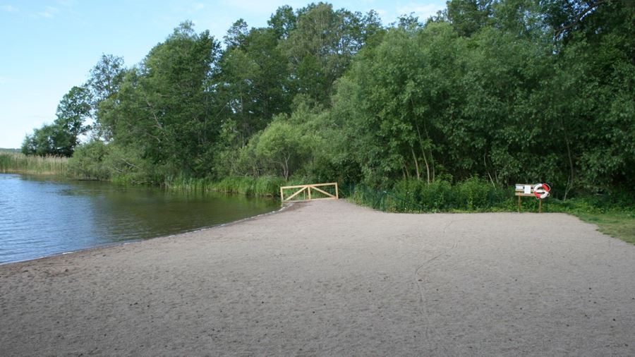 Lerhagsbadet hundbad, stranden med staket och grind. En livboj finns upphängd vid en anslagstavla på stranden.