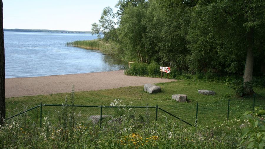 Hundbadets stranden och gräsmatta syns innanför staketet. En anslagstavla med en livboj finns på stranden.