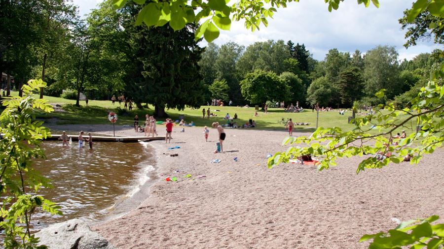 Ett femtiotal personer befinner sig på gräsmattan, stranden eller i vattnet vid Lyssnaängsbadet.