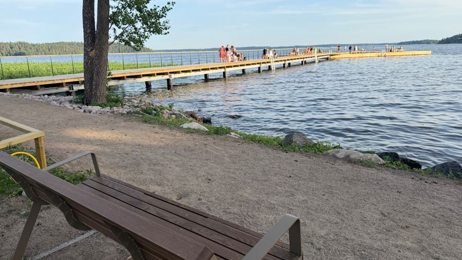 En bänk vid strandkanten och utsikt över brygga och vatten