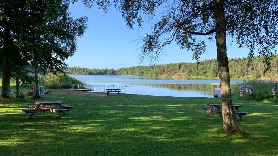 Södersjöbadet med bänkar med bord, soptunnor, brygga och tillgänglighetsanpassad badramp till vattnet. Fotografi.