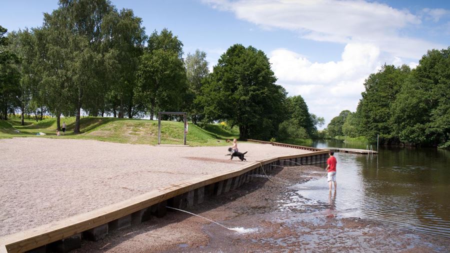 Ett barn och en vuxen leker med en hund på gruset vid Storvadsbadet.