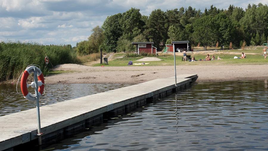 Några personer sitter på gräset vid Testenbadet. En ser vass, en brygga och ett vindskydd i trä.