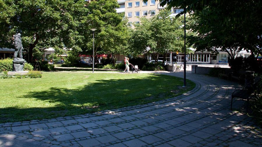 En kvinna och en man med barnvagn går förbi en stenstaty föreställande en man i Börjeparken/Finn Malmgrens plan.