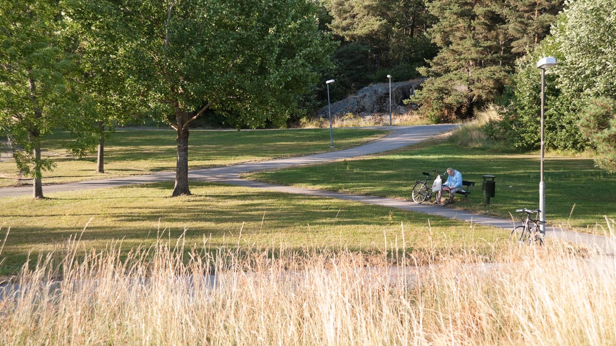 En man sitter på en parkbänk längst en gång- och cykelbana som leder mellan gräsytor och träd i Flogstaparken.