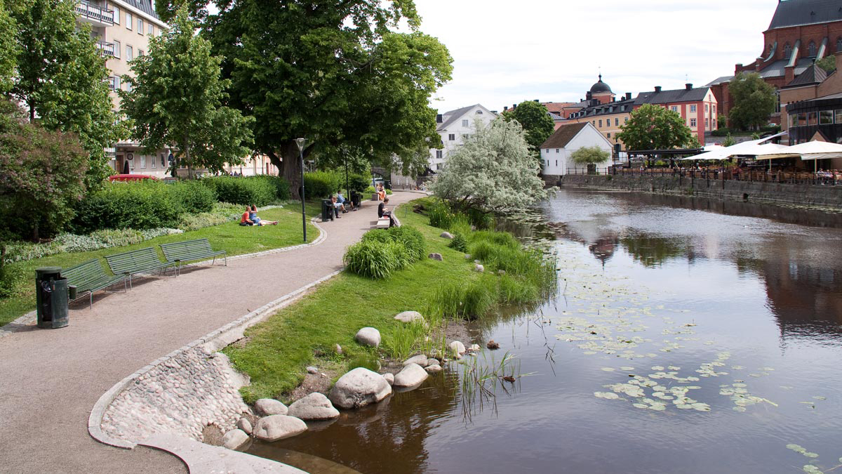 Några personer sitter på gräsytor och bänkar vid en grusväg i Gotlandsparken som ligger jämte Fyrisån.