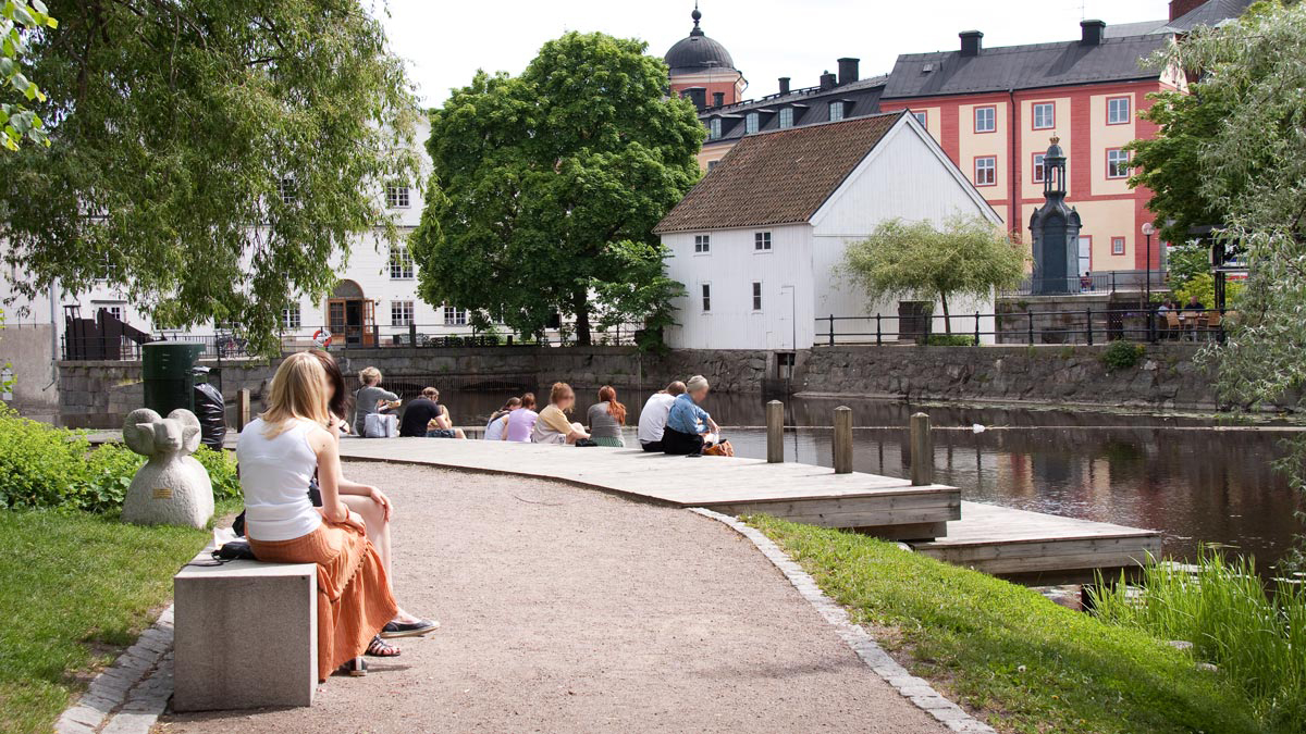 Ett tiotal personer sitter på en bänk eller på en brygga intill Fyrisån i Gotlandsparken.