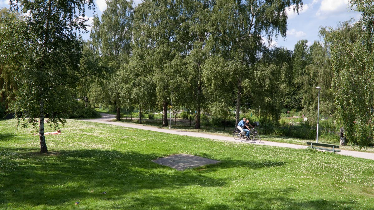 Två män cyklar på en gång- och cykelväg mellan gräsytor och träd på Höganäshöjden. En person ligger och solar i gräset.