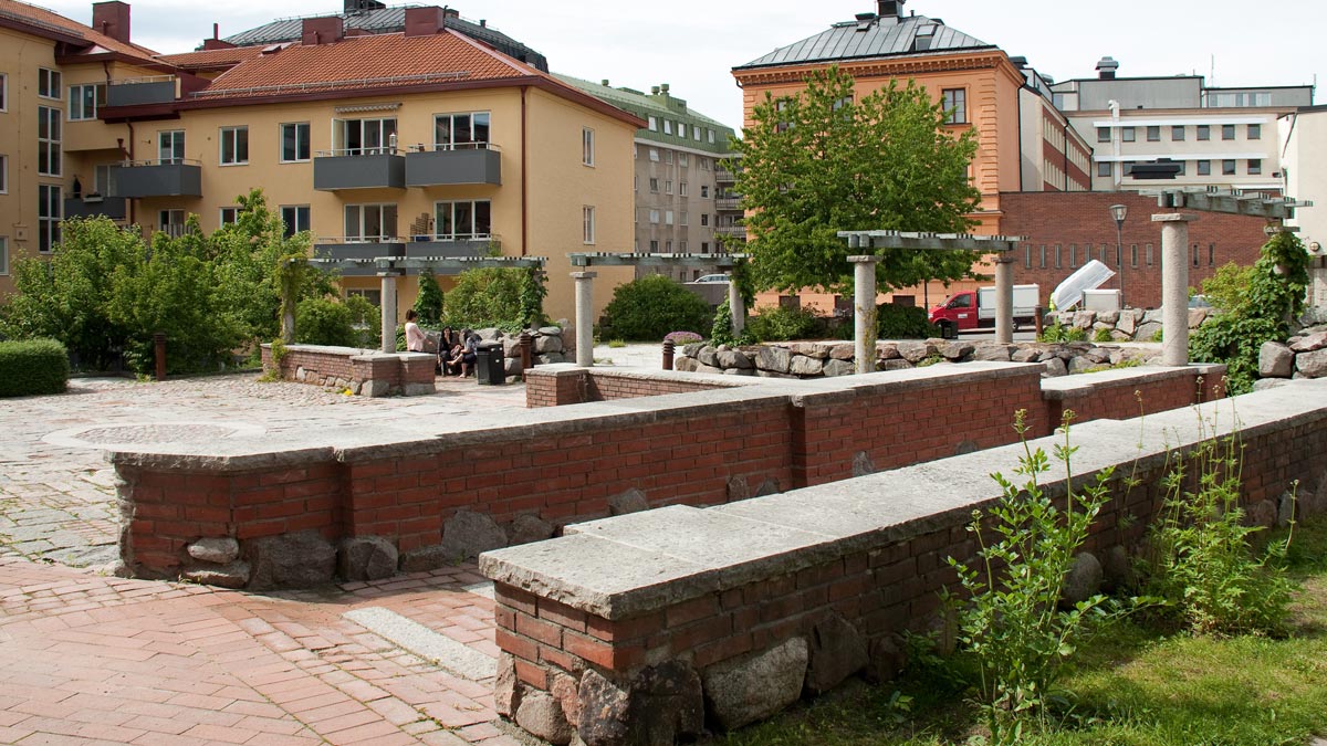Några personer sitter på en bänk intill tegelstensmurar i Klosterparken.