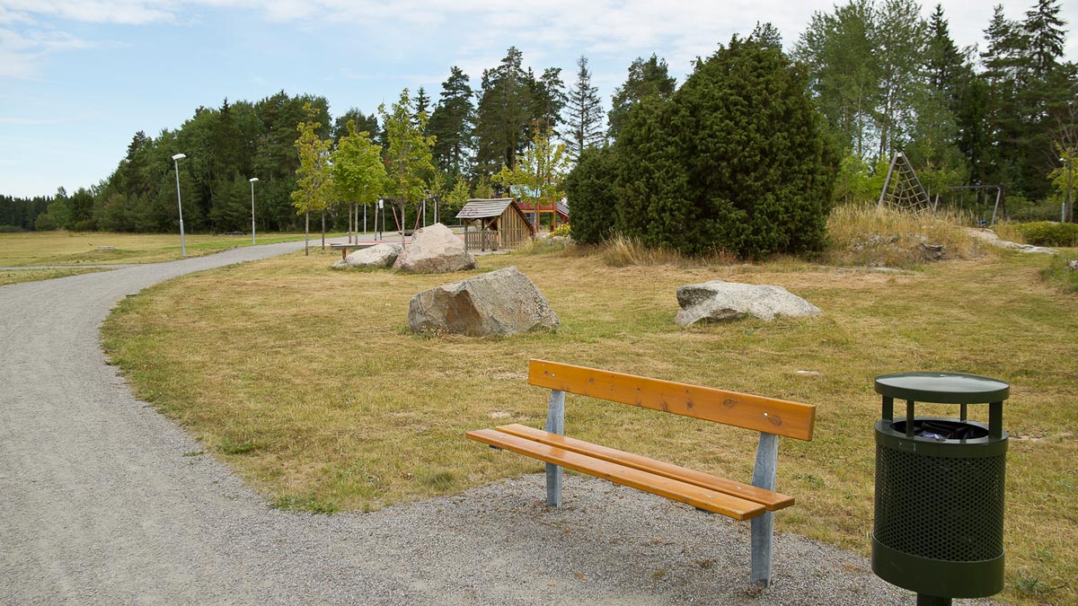 En grusväg går förbi gräsytor med stora stenar, träd och en lekplats. En bänk står längs vägen.