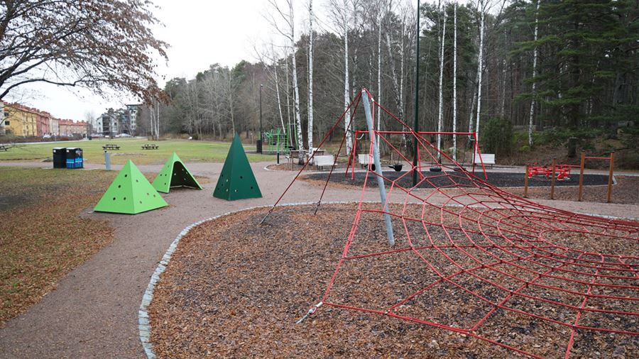 I förgrunden syns ett rött klätternät. Bakom det syns tre gröna, trekantiga hyddor att gå in i. De har runda hå i väggarna som man kan se ut genom. I bakgrunden syns en gungställning och utegymmet.