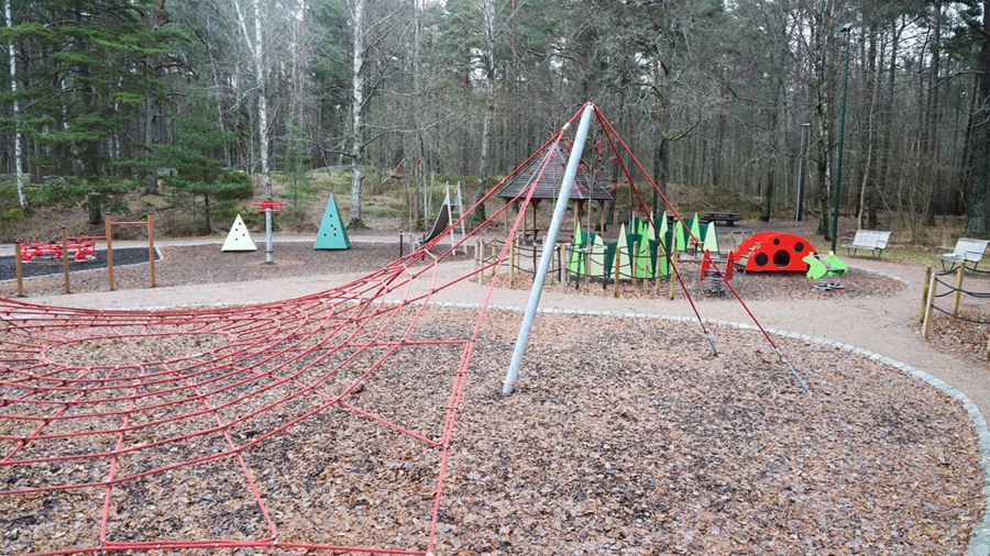 Ett rött klätternät syns i förgrunden. Bakom det syns en picknickplats med tak, en gunga som ser ut som en groda och annan lekutrustning.