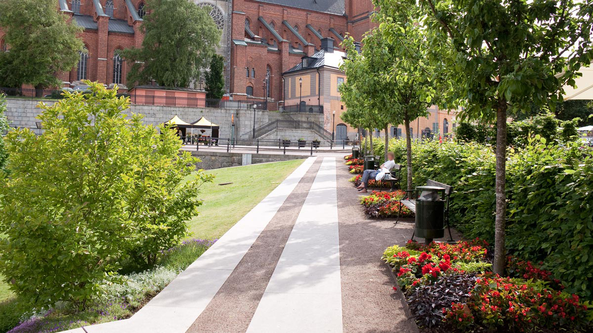 En rak gång mellan en gräsplätt, bänkar och blomsterodlingar. Uppsala domkyrka syns i bakgrunden.