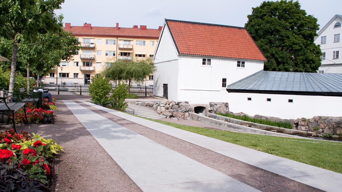 En sten- och grusgång med bänkar, gräs och blomsterodlingar på sidorna. Ett vitt trähus till höger.