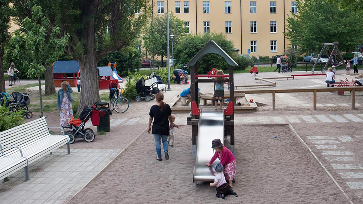 Ett tiotal barn och vuxna på en lekplats med bland annat en klätterställning, gungor och en sandlåda.
